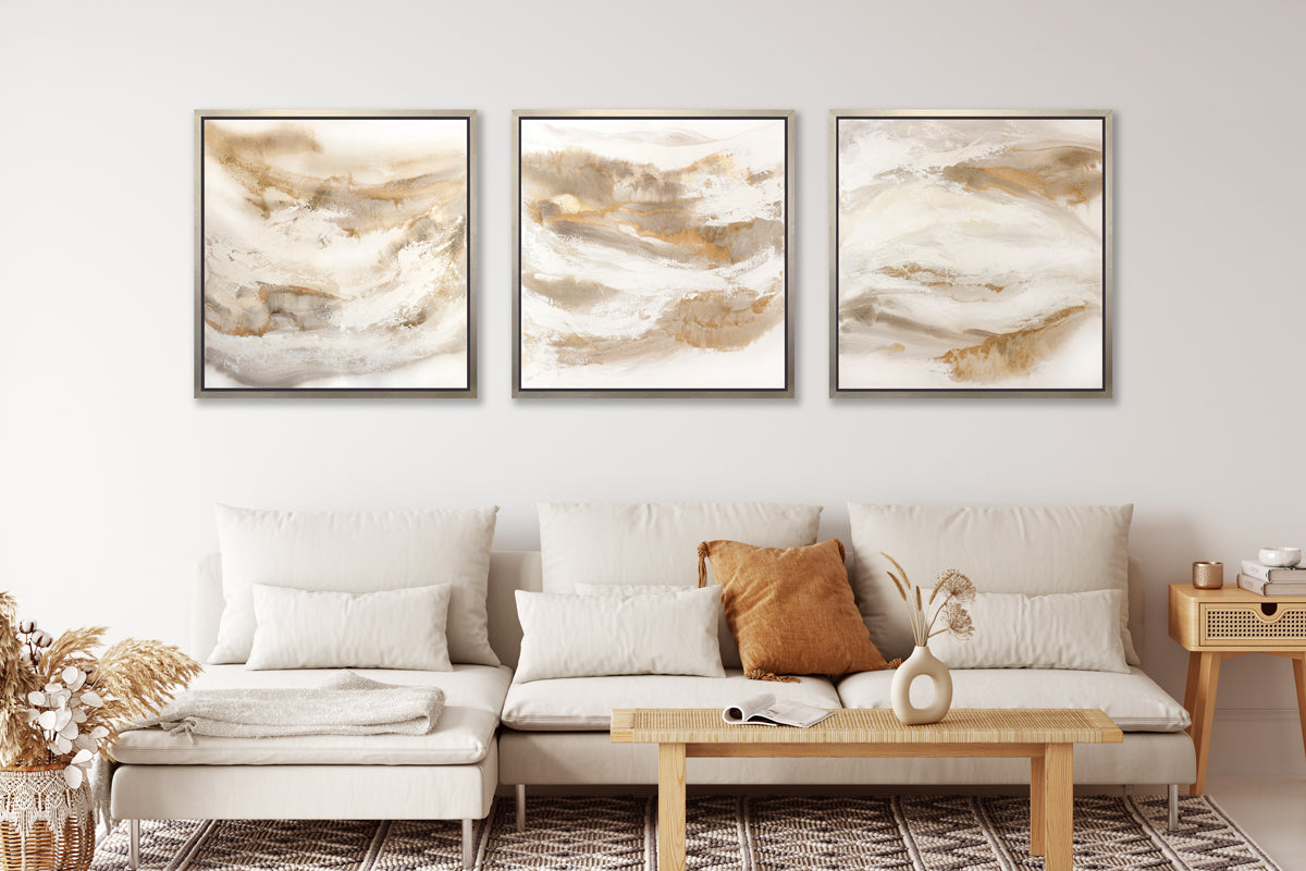 Three gold, silver, white and beige abstract prints in silver floater frames hang side by side on a white wall above a white sofa with pillows, a rattan coffee table with a white vase and a wood side table with a stack of books and a copper container.