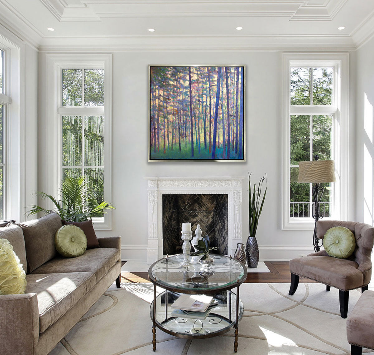 A colorful tree landscape in a silver floater frame hangs on a white wall above a fireplace across from a tan sofa with green pillows, two chairs with green pillows and a oval glass coffee table.