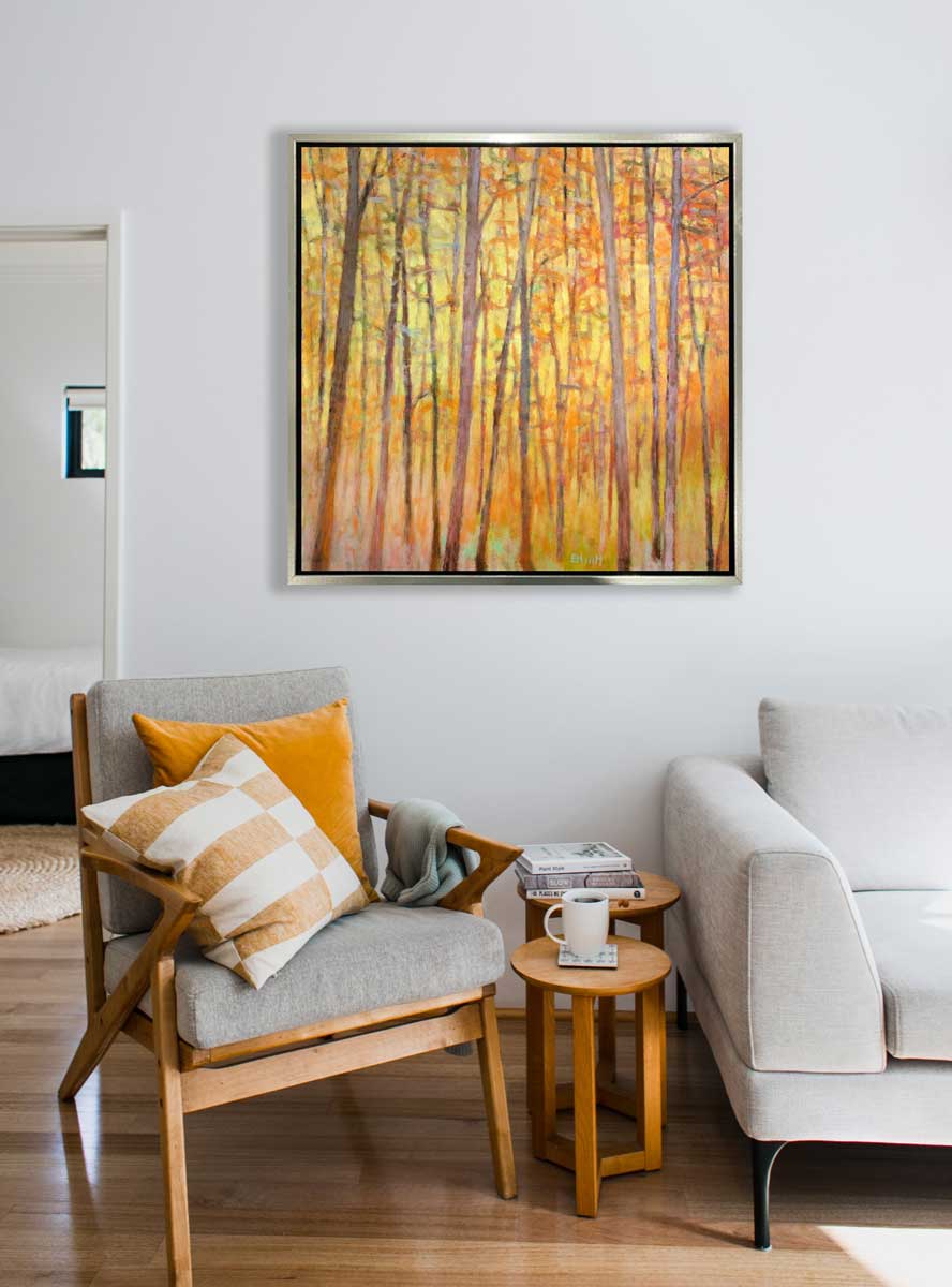 A yellow and orange impressionistic forest print in a silver floater frame hangs on a white wall above a mid century chair with light grey cushions and orange throw pillows.  