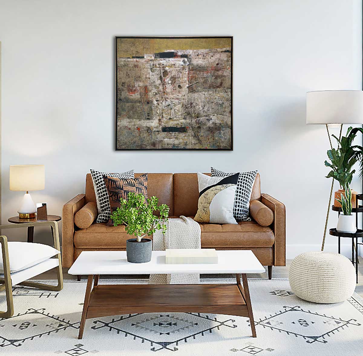 A white and grey abstract painting in a gold floater frame hangs on a white wall above a brown leather sofa with printed pillows, a white marble and wood coffee table and a tall floor lamp.