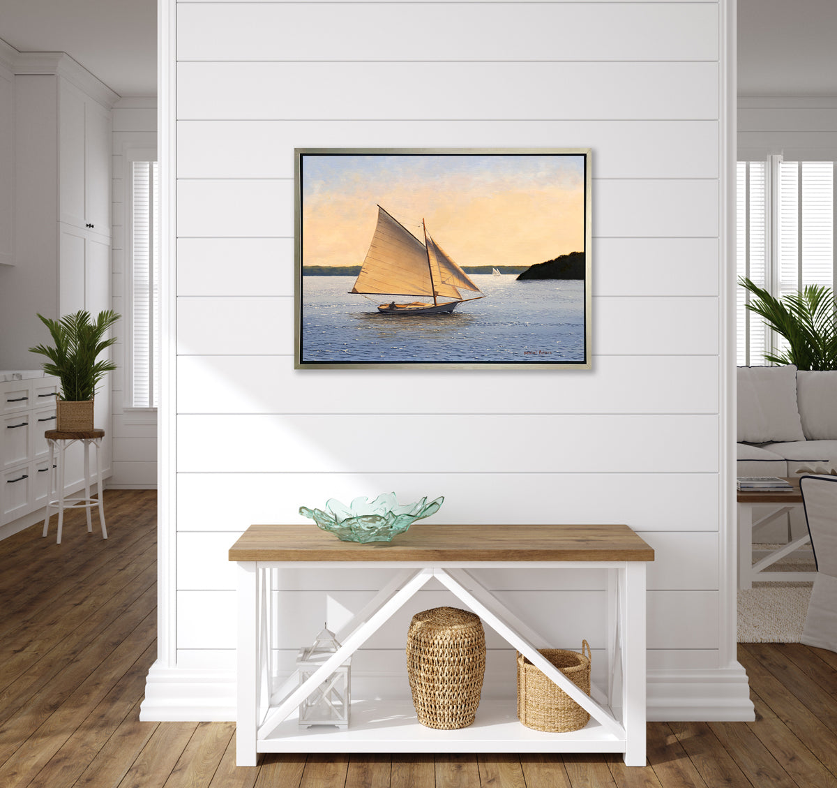 A blue, beige and green seascape print with sailboats in a silver floater frame hangs on a white wall above a white console table with a teal decorative glass bowl, two woven baskets and a white lantern.