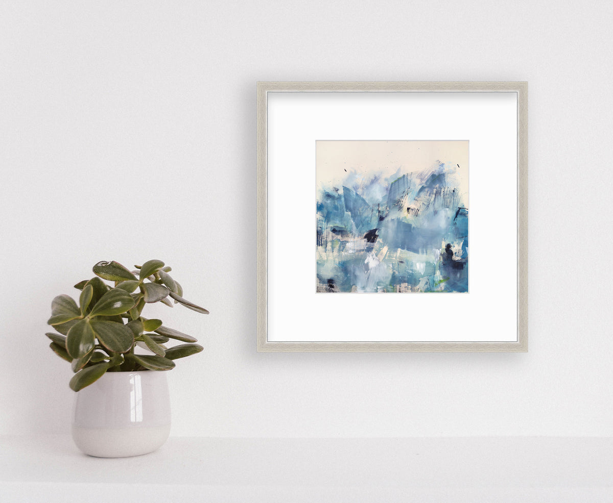 A blue paper print matted and framed in a silver frame hangs on a white wall above a small plant in a white planter. 