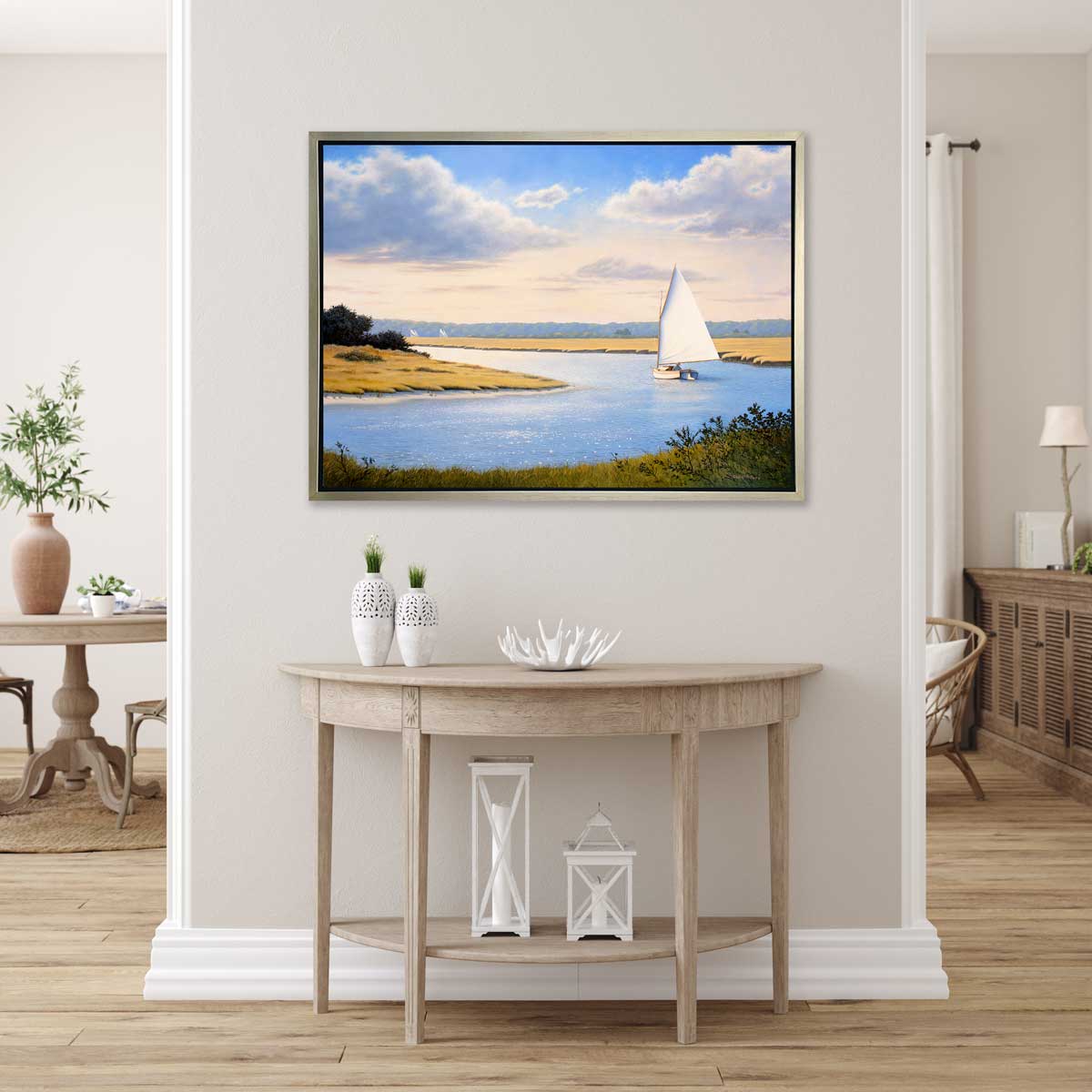 A print of a sailboat going down a winding blue river in a silver floater frame hangs on a white wall above a natural wood half-circle console table with two white vases, a white decorative bowl and two white lanterns.