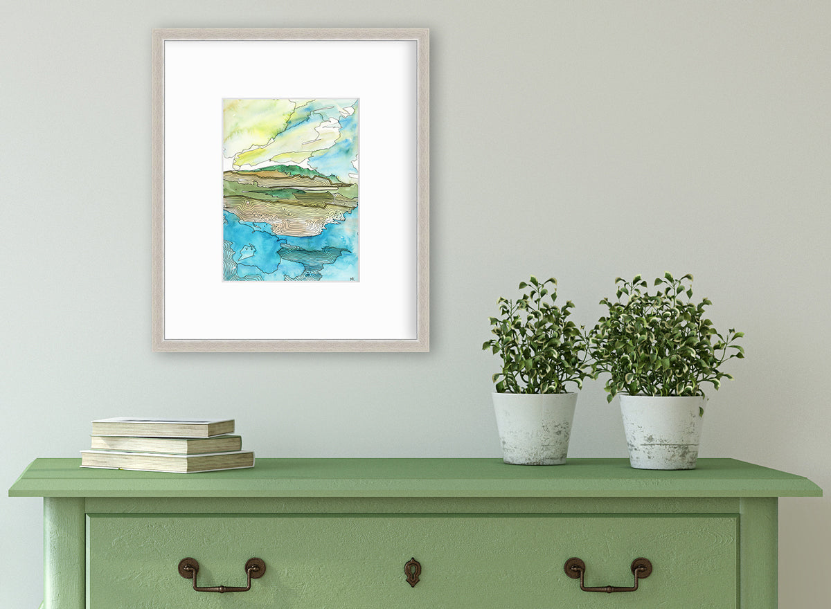 A blue, green, brown and light yellow abstract landscape with black outlines print in a silver frame with a mat hangs on a white wall above a green console table with a stack of books and two potted plants.