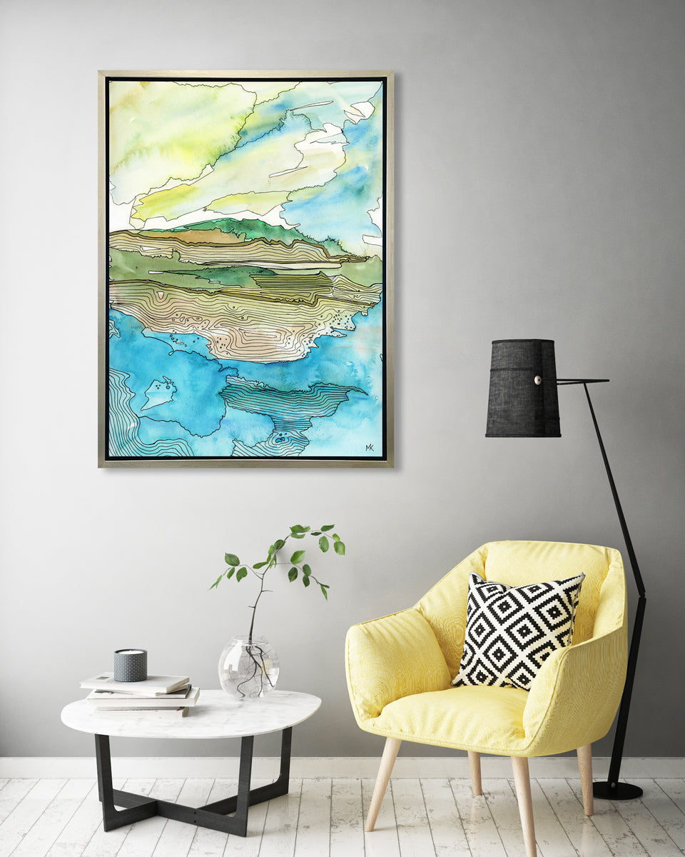 A blue, green, brown and light yellow abstract landscape with black outlines print in a silver floater frame hangs on a white wall above a yellow chair with a black and white printed pillow, a short round side table with a stack of books and a clear glass vase with a greenery stem and a black floor lamp.