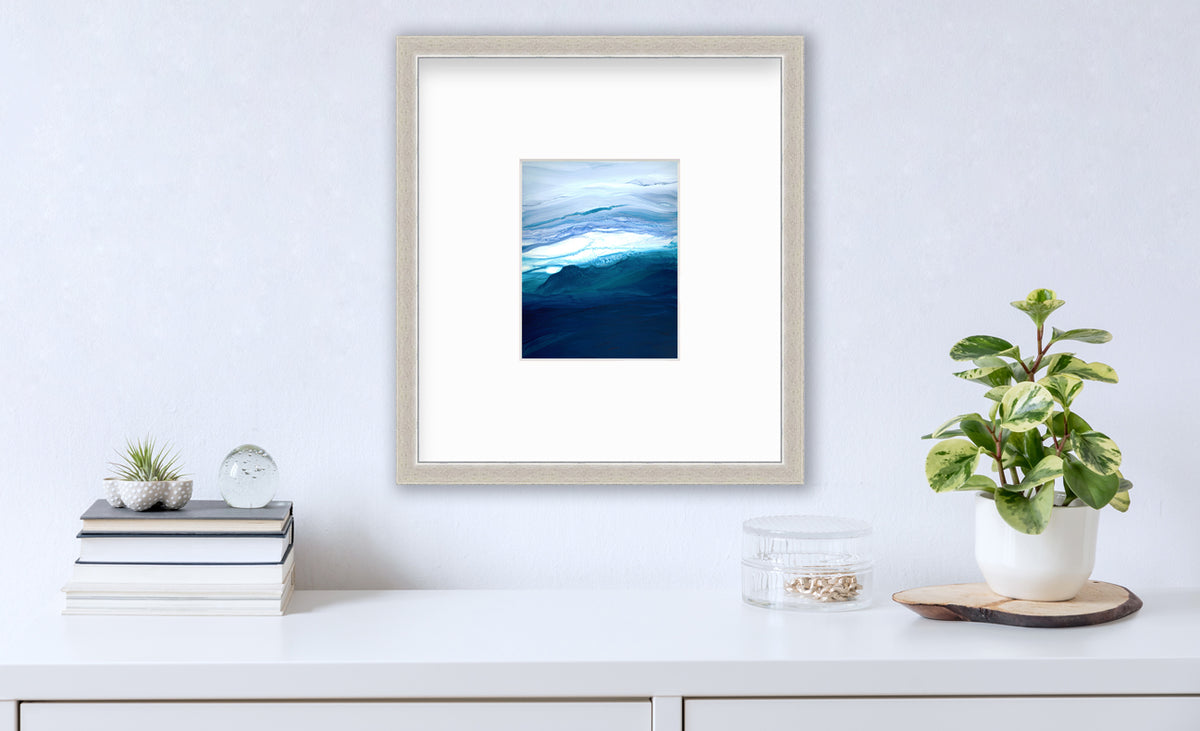 A blue, teal, lavender and white abstract print in a silver frame with a mat hangs on a white wall above a white surface with a stack of books, a round clear container with gold accessories and a potted plant.
