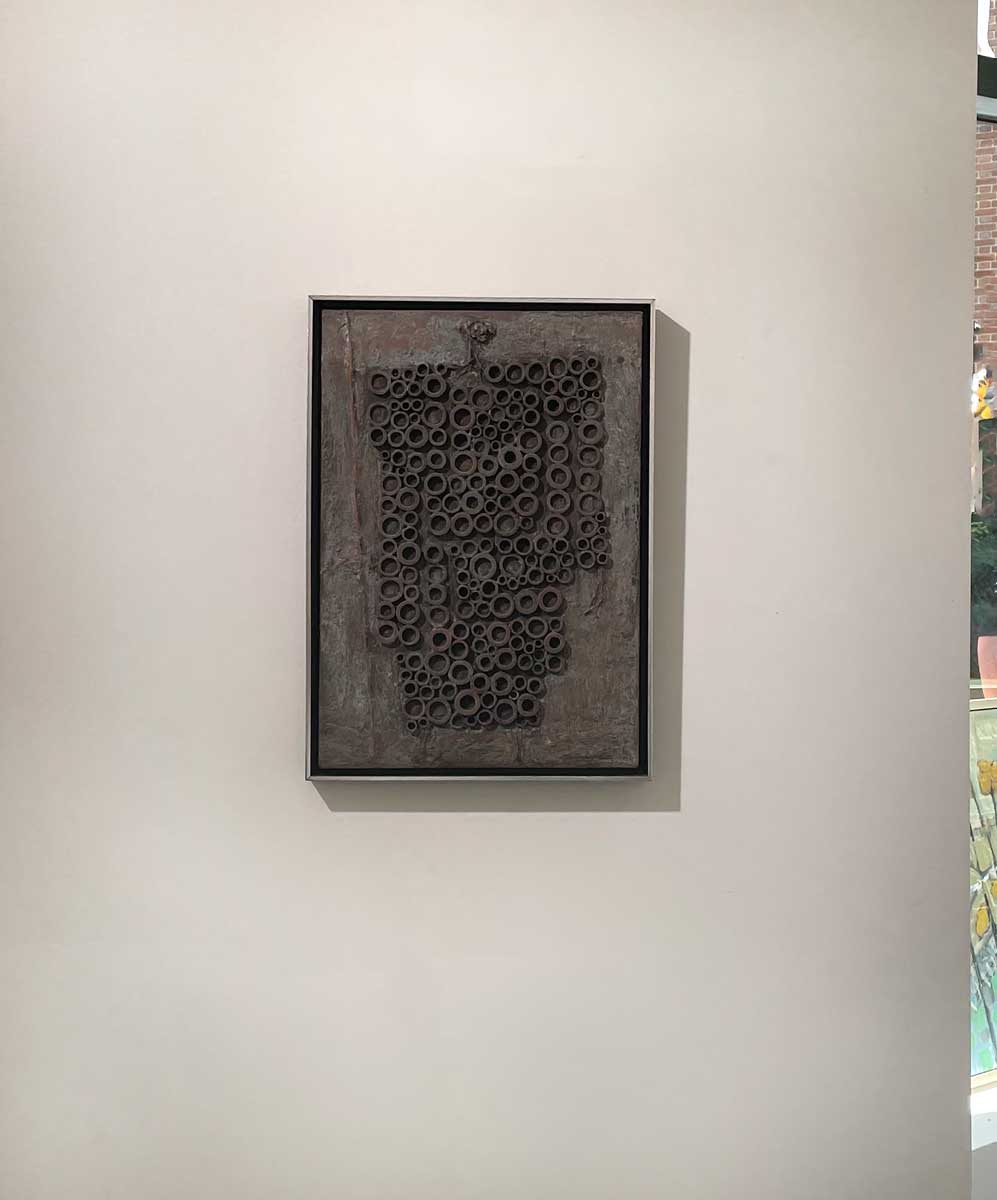 A grey abstract painting with embedded wood circles in a silver floater frame hangs on a white wall.