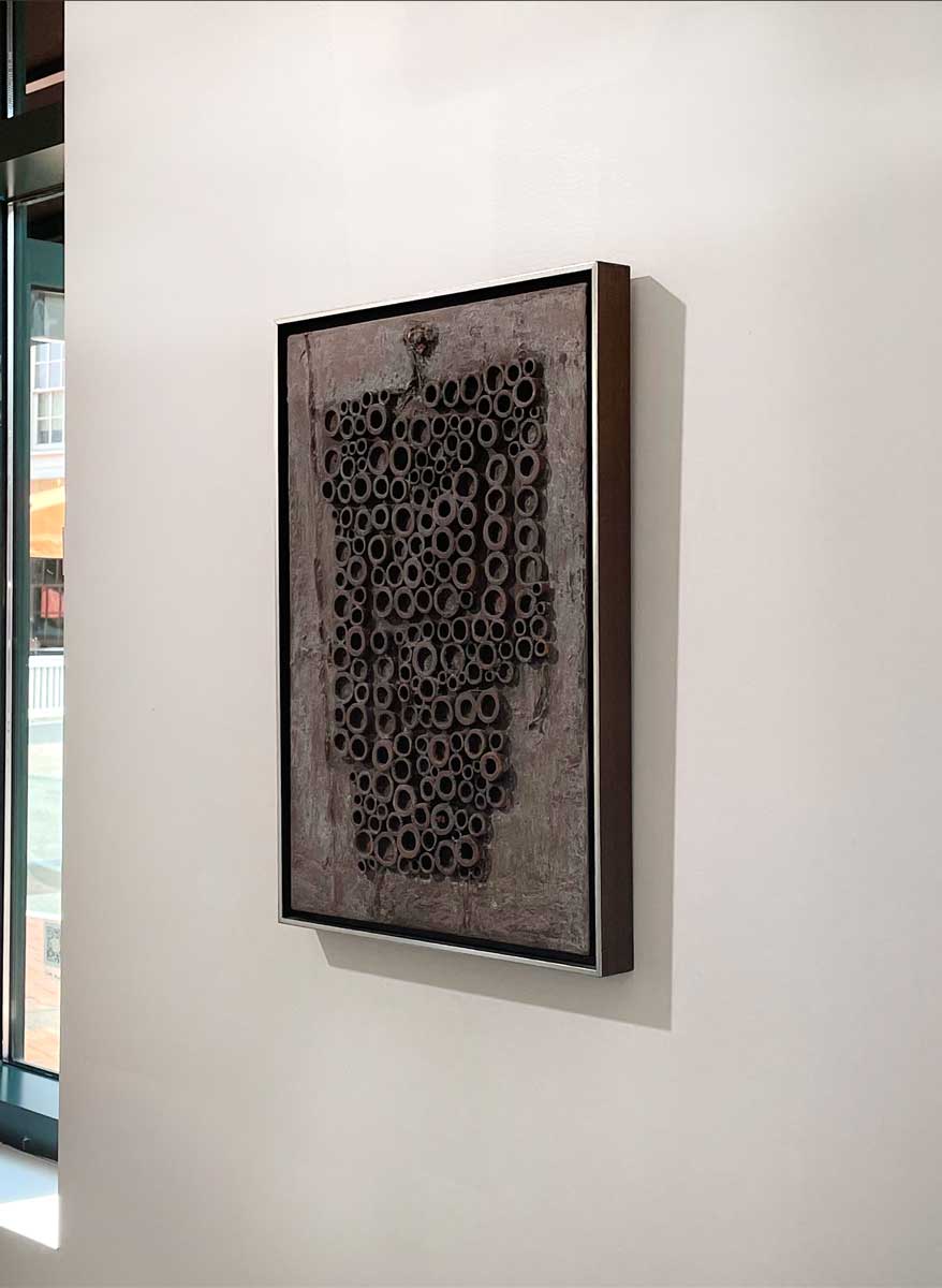 A right side-angle view of a grey abstract painting with embedded wood circles in a silver floater frame hangs on a white wall.