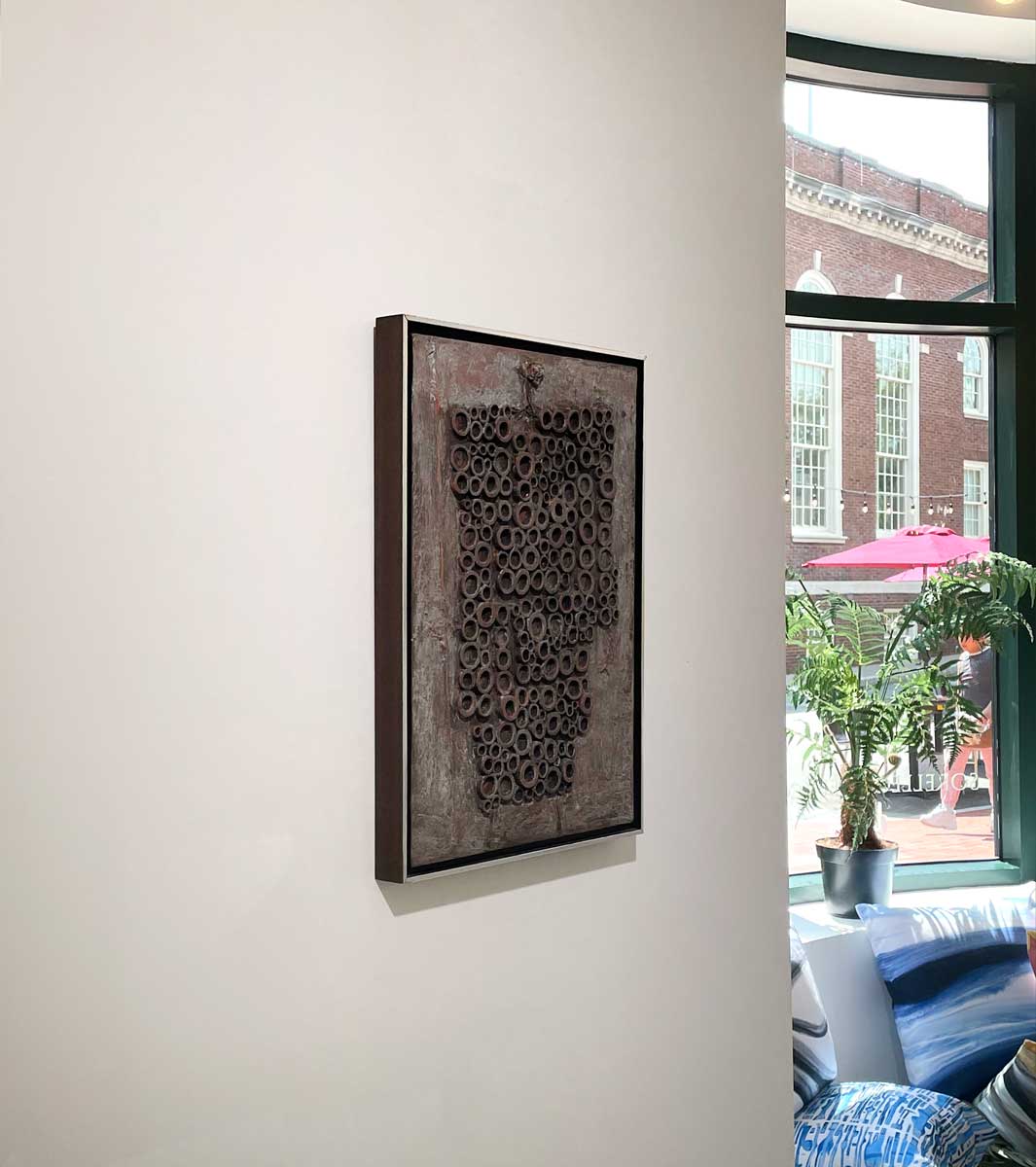 A left side-angle view of a grey abstract painting with embedded wood circles in a silver floater frame hangs on a white wall.
