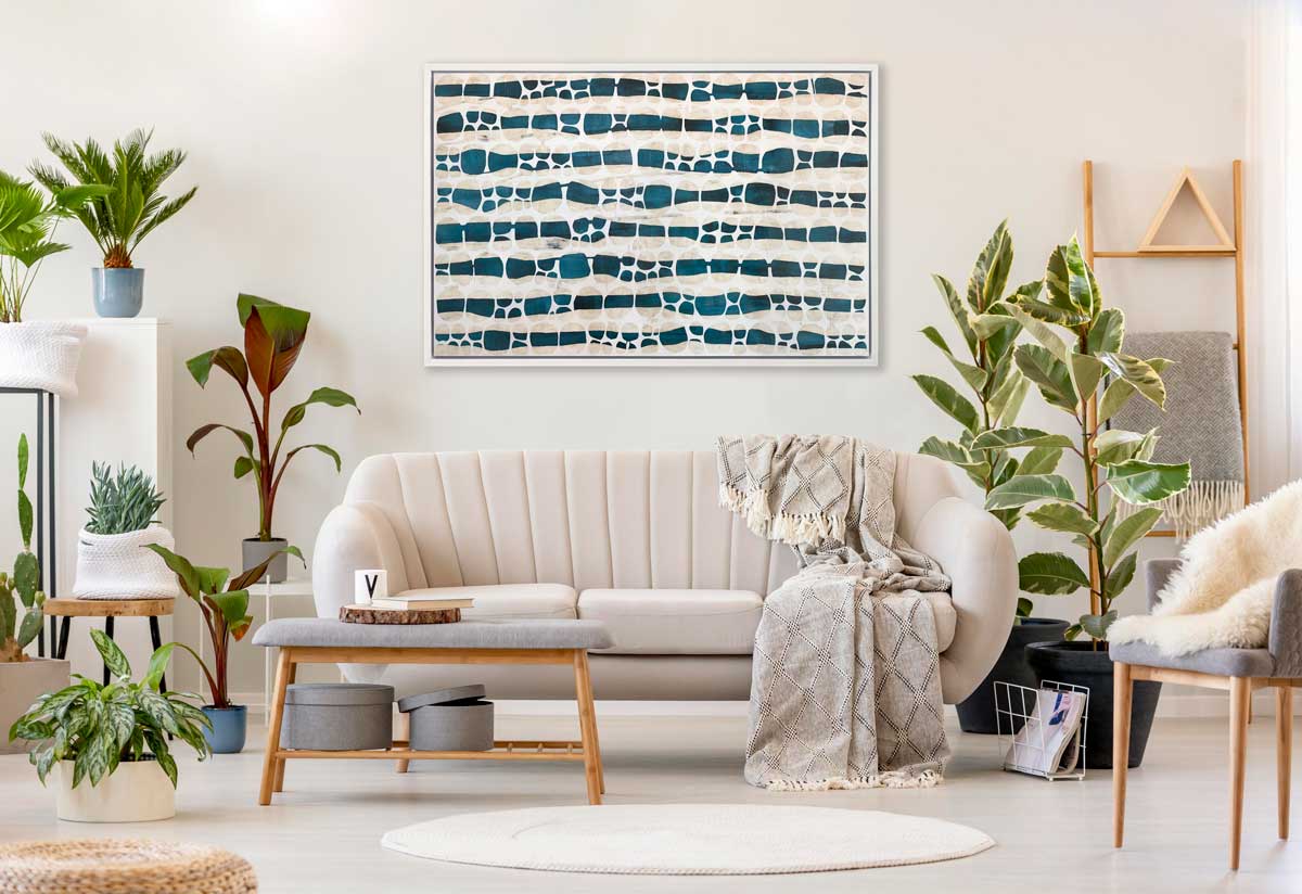 A white, dark teal and beige abstract print in a white floater frame hangs on a white wall above a white sofa with a throw blanket, several potted floor plants and a grey bench.