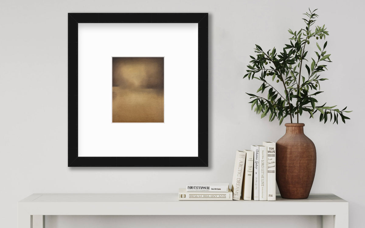 A brown and gold abstract print in a black frame with a mat hangs on a white wall above a white console table with a stack of books and a terracotta vase filled with greenery stems.