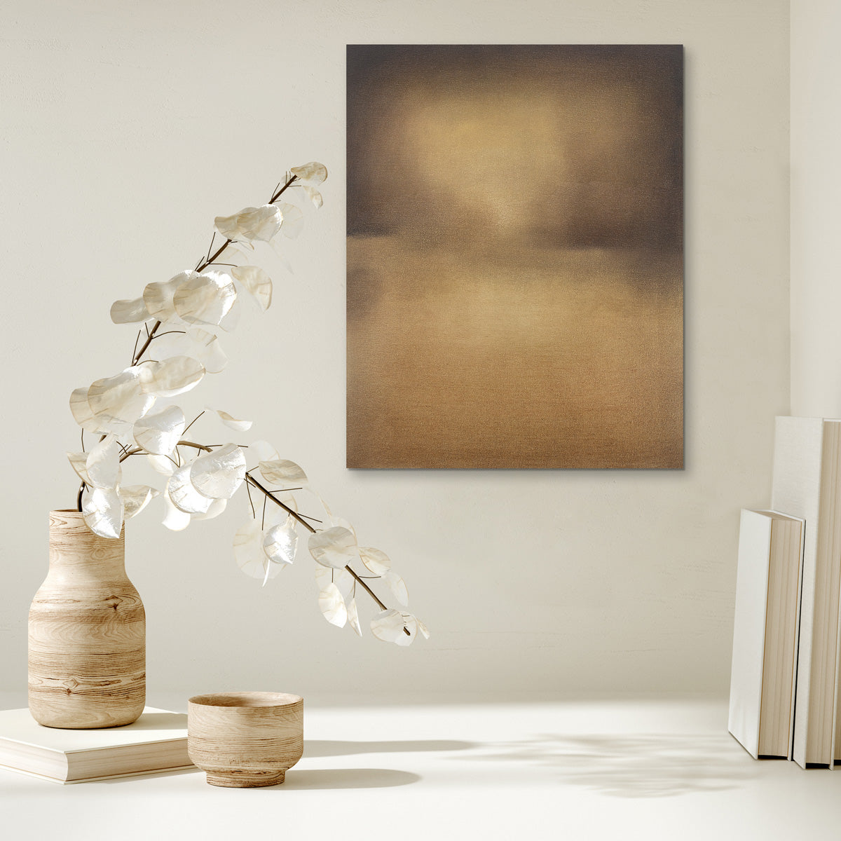 A brown, beige and goldenrod abstract painting by Tony Iadicicco hangs on a white wall above a stack of books and a wooden vase with white leaf stems.