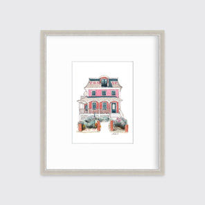 A pink and red house print in a silver frame with a mat hangs on a white wall.