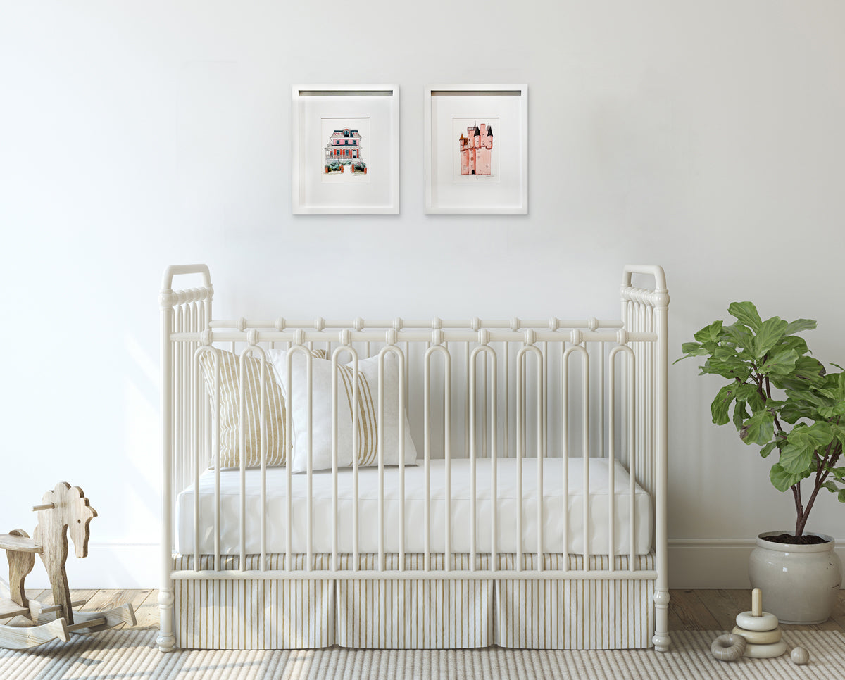 Two architectural illustration drawings in white frames with mats hang on a white wall above a white iron crib with neutral bedding, a wooden rocking horse and a potted floor plant. 