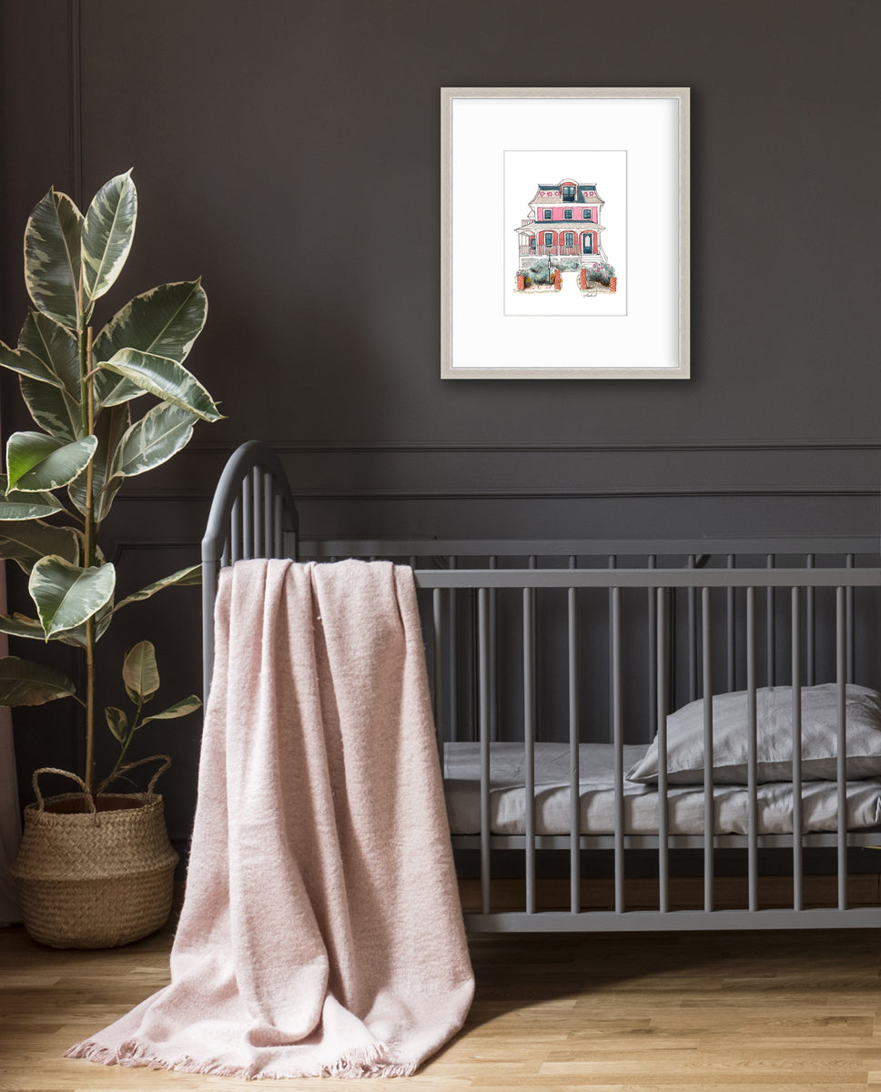 A pink and red house print in a silver frame with a mat hangs on a dark grey wall above a grey crib with grey bedding, a light pink throw blanket over the side and a large potted floor plant.