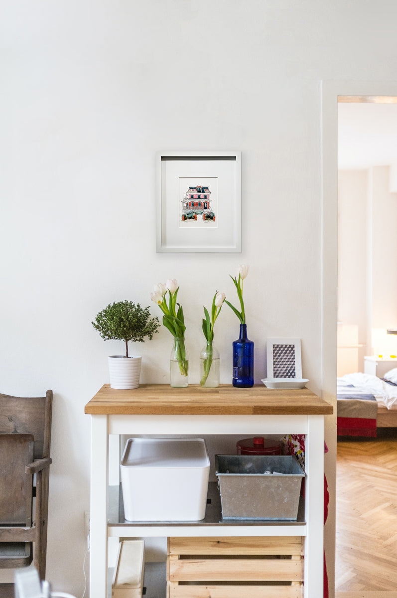 A pink, beige, red and slate blue illustration drawing of a house in a white frame with a mat hangs on a white wall above a wood surface with a few bottled white tulips, a potted shrub and a white dish.