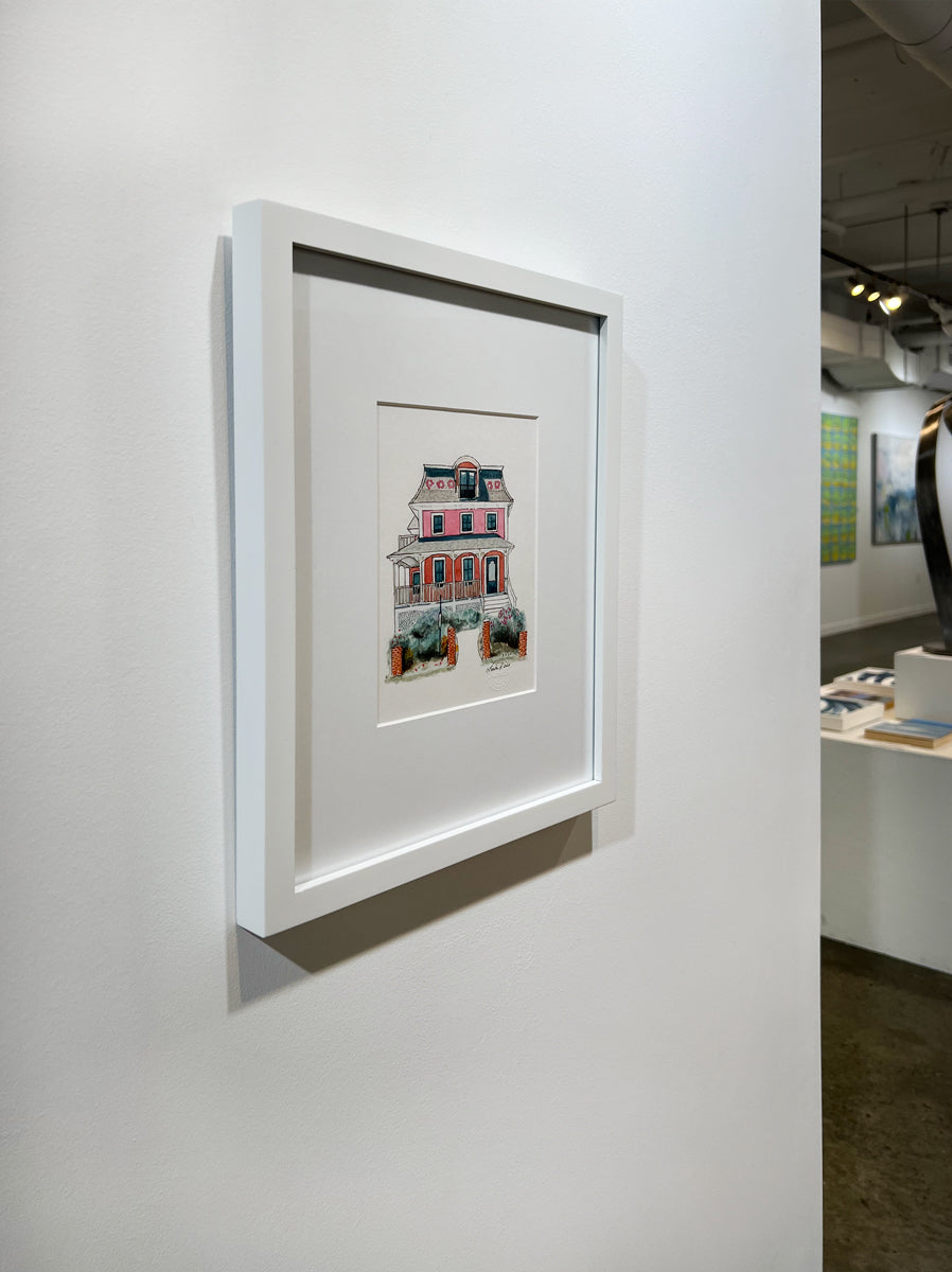 A left side-angle view of a pink, beige, red and slate blue illustration drawing of a house in a white frame hangs on a white wall.
