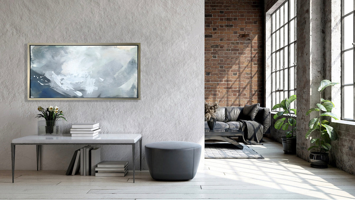 A blue, grey and white abstract print in a silver floater frame hangs on a grey wall above a white table with a stack of books, a potted floral arrangement and a grey ottoman.
