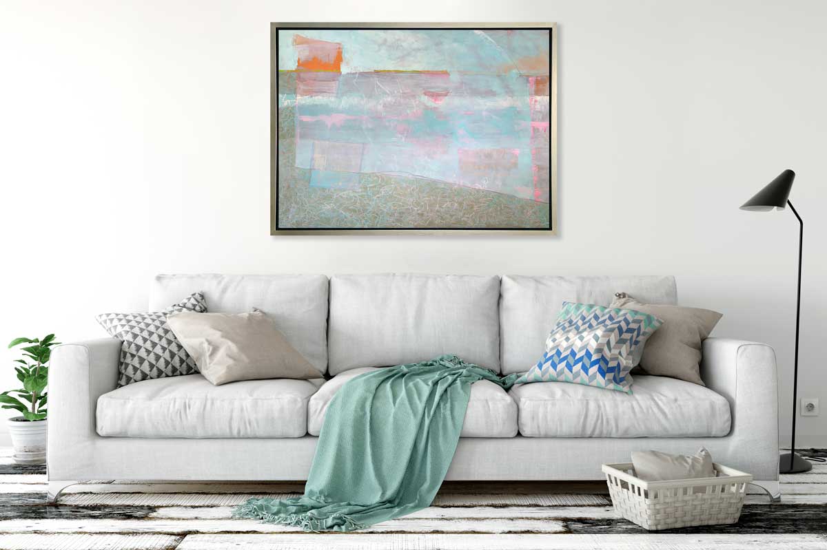 A light blue and pink abstract print in a silver floater frame hangs on a white wall above a grey sofa with pillows and a teal throw blanket, a small white basket with a pillow and a black floor lamp.