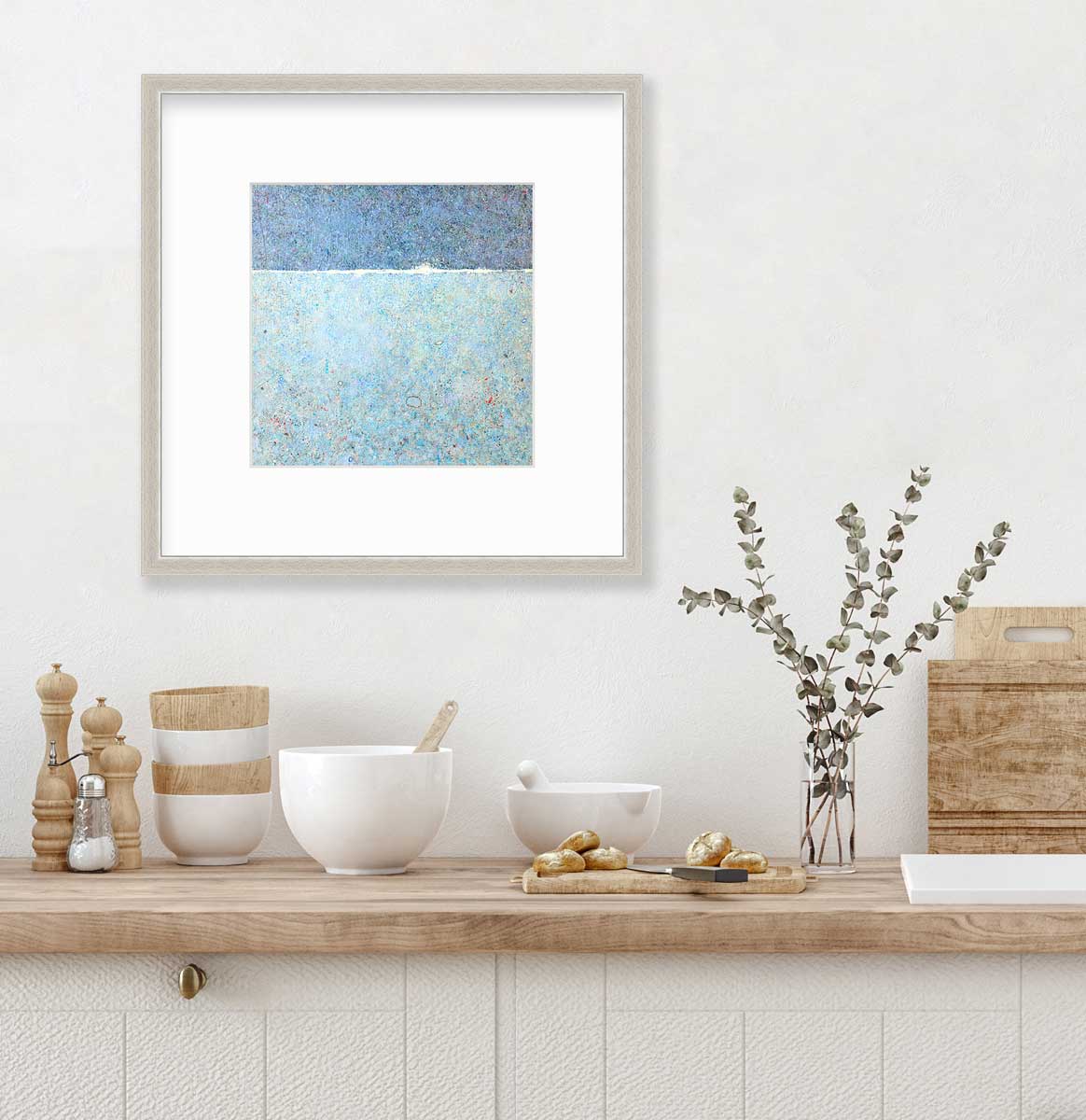 A blue abstract landscape print in a silver frame with a mat hangs on a white wall above a butcher block countertop with bowls, a cutting board and a clear glass vase with eucalyptus stems.
