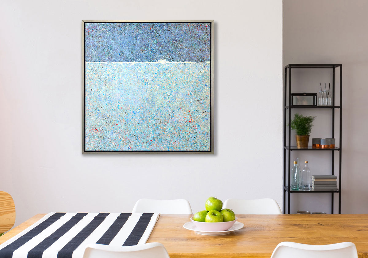 A blue abstract landscape print in a silver floater frame hangs on a white wall above a wooden table with a blue and white striped table runner, a bowl of green apples and four white chairs.