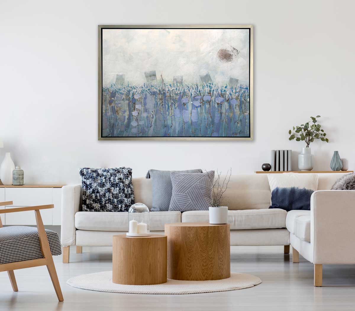 A beige, teal and lavender abstract print in a silver floater frame hangs on a white wall above a white sectional sofa with blue pillows, a small shelf with books and vases and two wood round coffee tables.