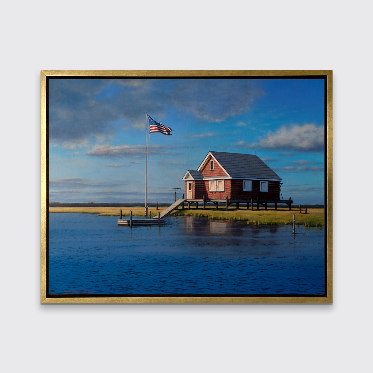 A print of a red bay house looking over a body of water in a gold floater frame hangs on a white wall.