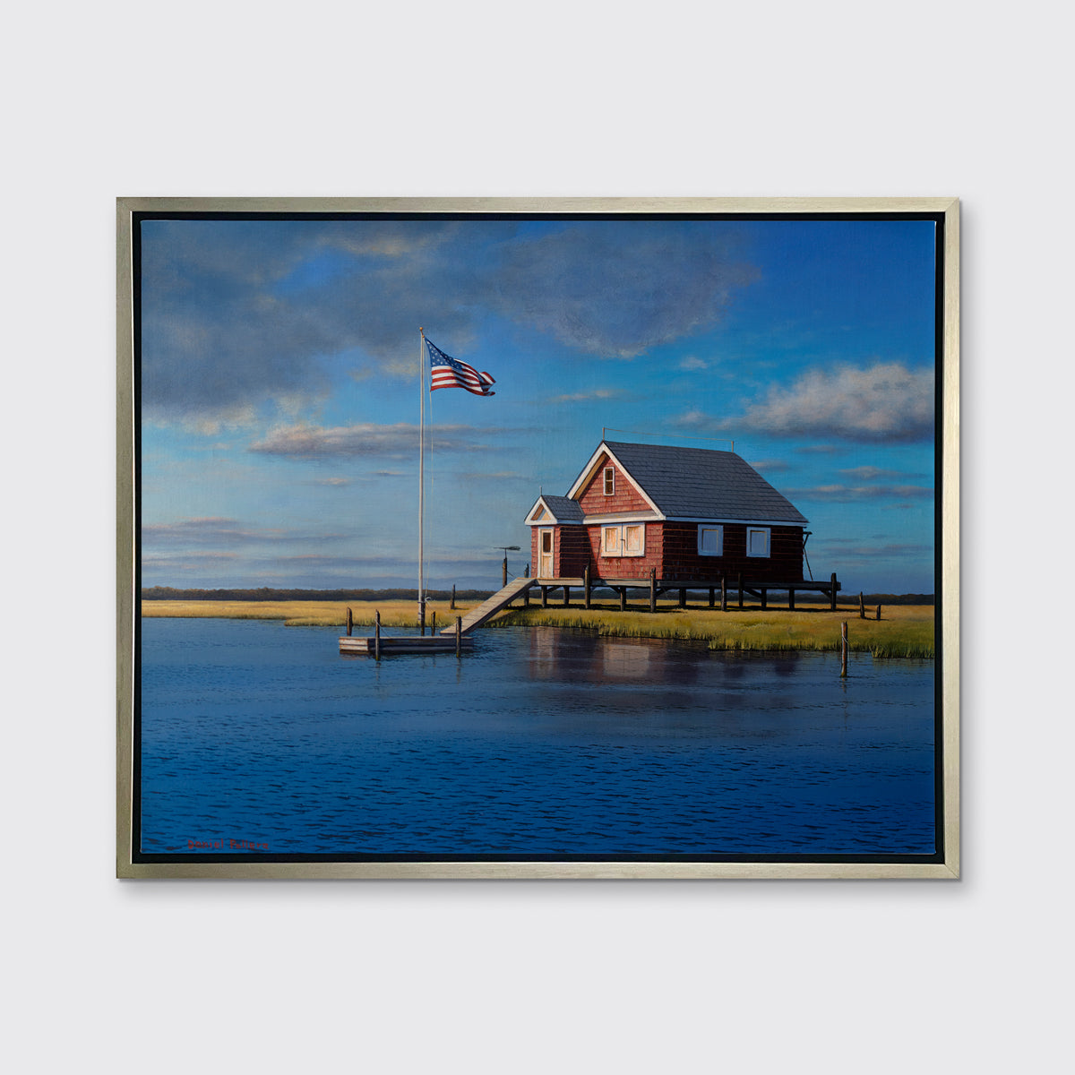A print of a red bay house looking over a body of water in a silver floater frame hangs on a white wall.
