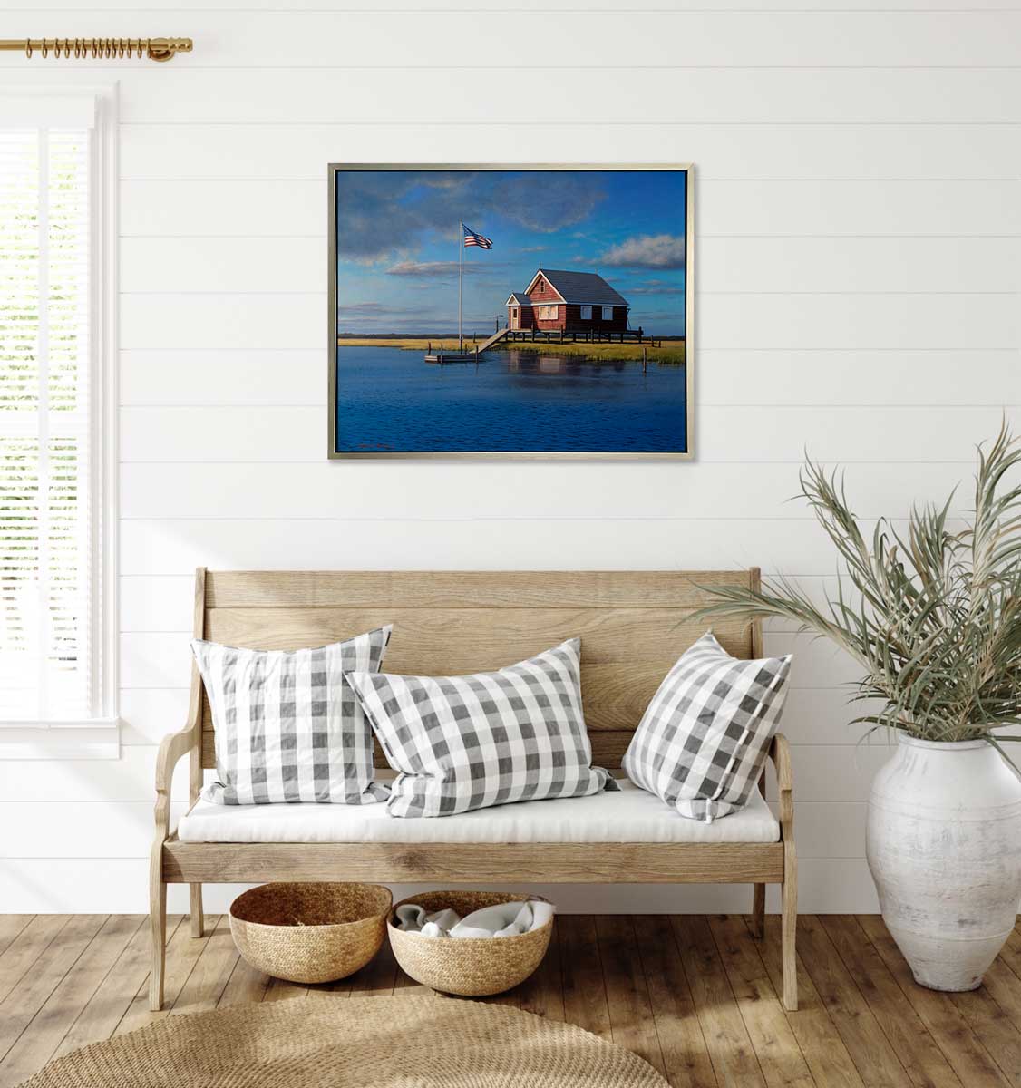 A print of a red bay house overlooking a body of water in a silver floater frame hangs on a white shiplap wall above a natural wood bench with white and grey checkered pillows, a white floor vase with grass stems and two woven baskets.