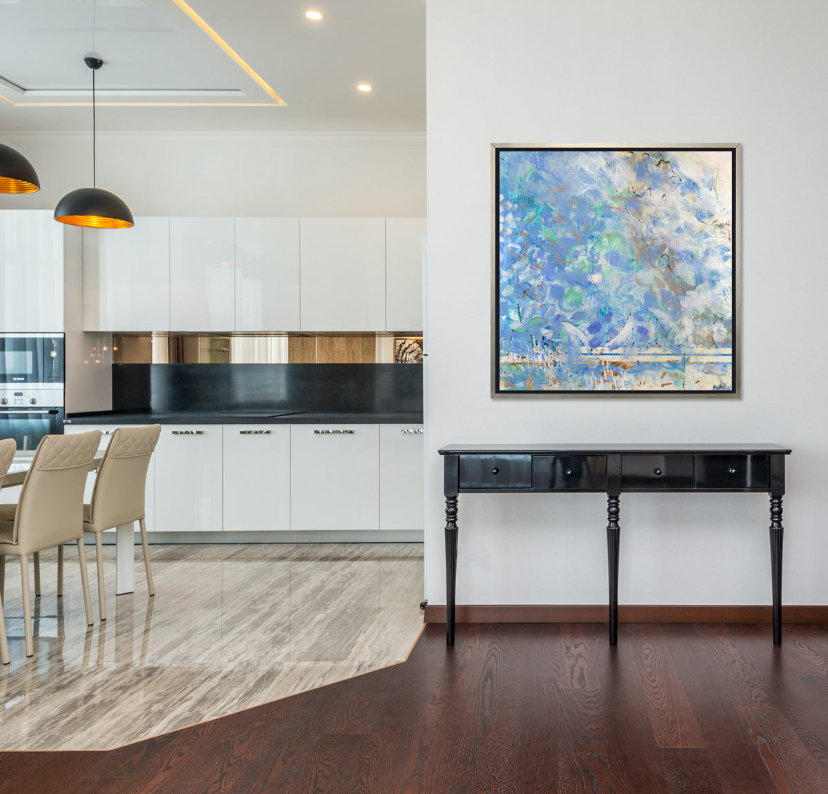 A lavender, blue and cream abstract print in a silver floater frame hangs on a white wall above a black console table and next to an opening for a kitchen with white cabinets, black countertops and a beige counter stools.