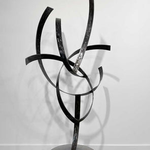 The back of abstract stainless steel sculpture with a grey metallic base sitting in front of a white wall.