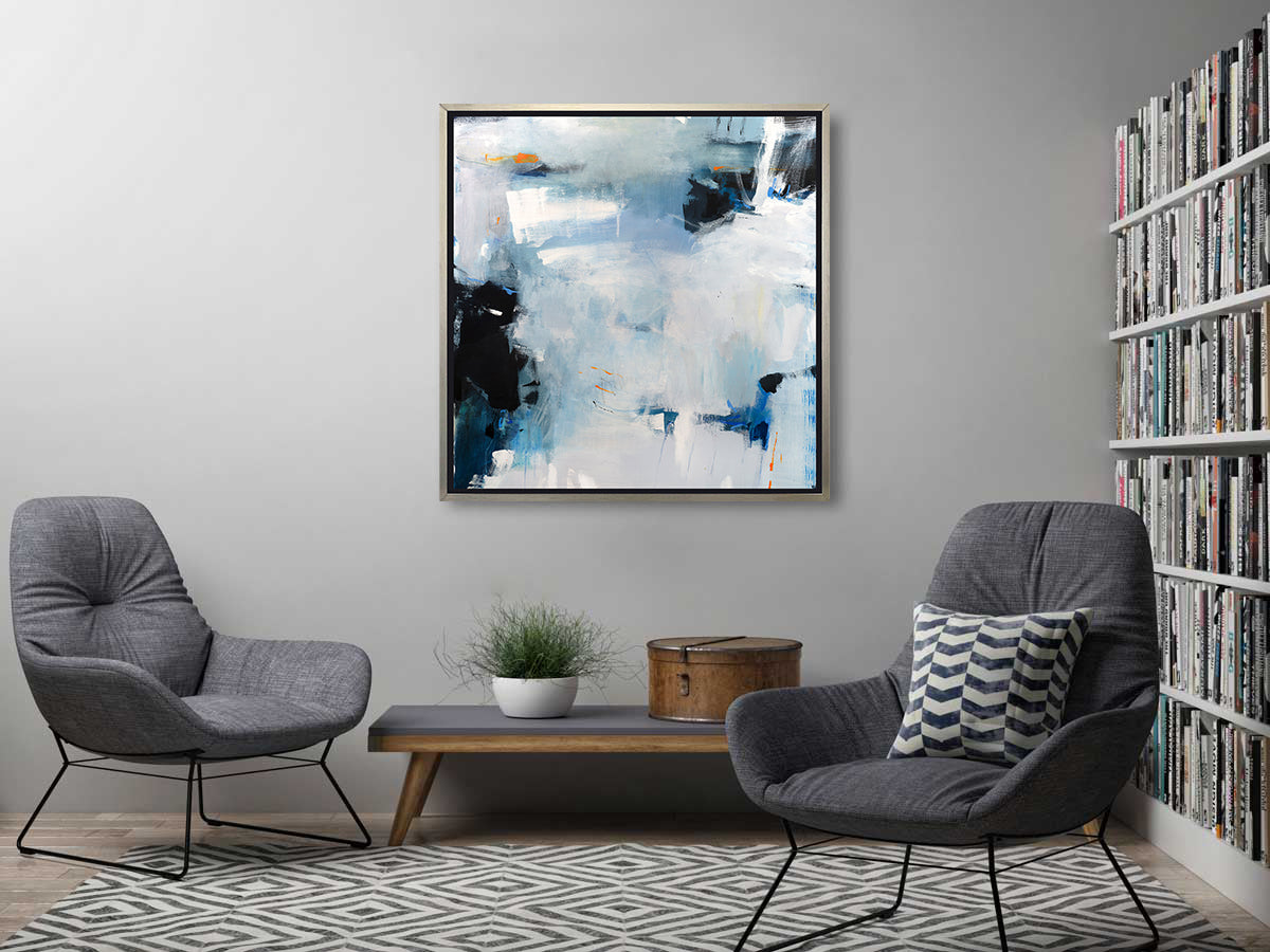 A blue and white abstract print in a silver floater frame hangs on a grey wall above a low wooden bench with a potted plant and a hat box, two grey chairs and a large bookshelf to one side.
