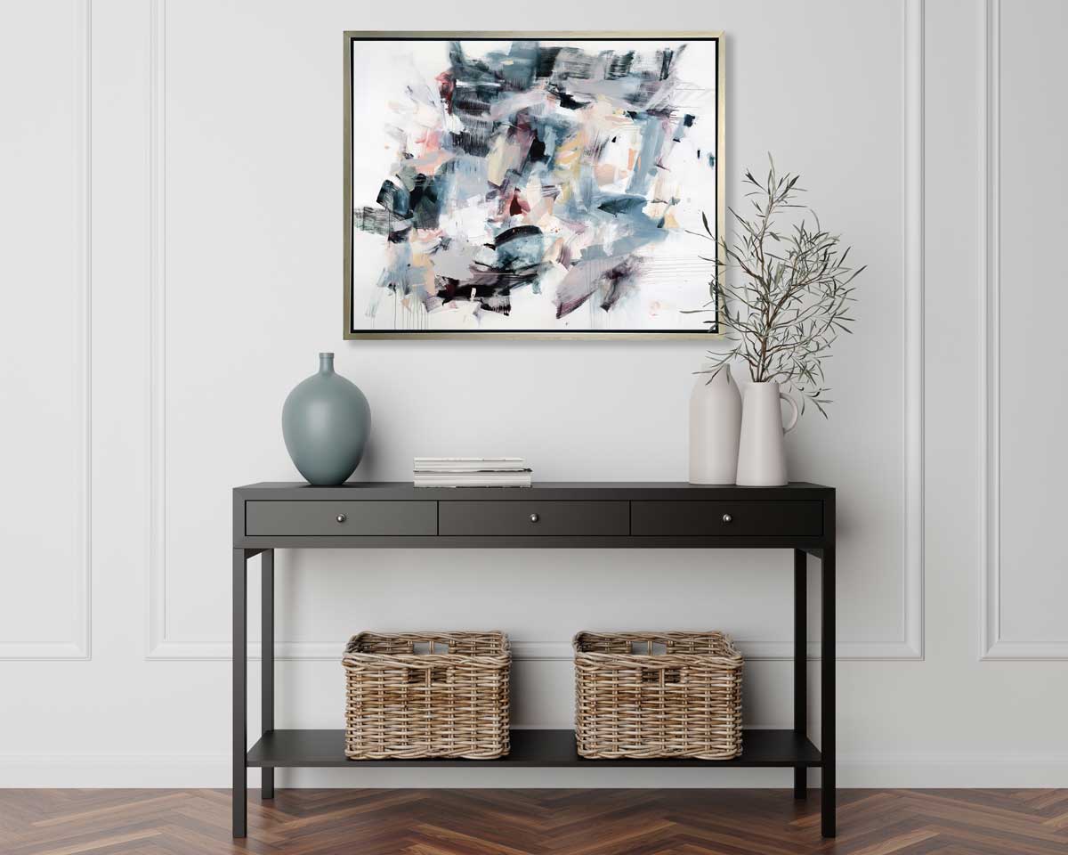 A multicolored abstract print in a silver floater frame hangs on a white wall above a black console table with a muted teal vase, two white vases with greenery and two woven baskets.