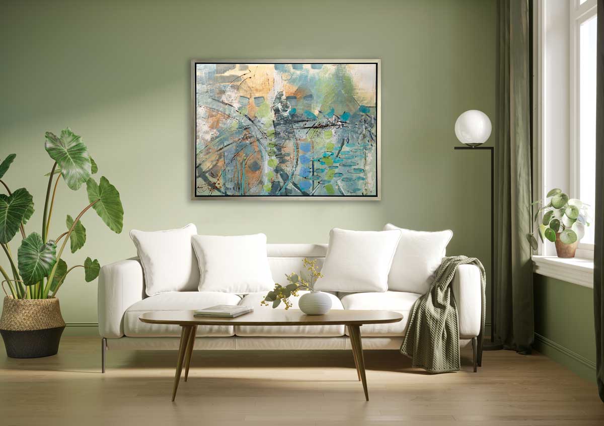 A blue, green, orange and black abstract print in a silver floater frame hangs on a light green wall above a white sofa with white pillows, a wood oval coffee table and a potted floor plant.