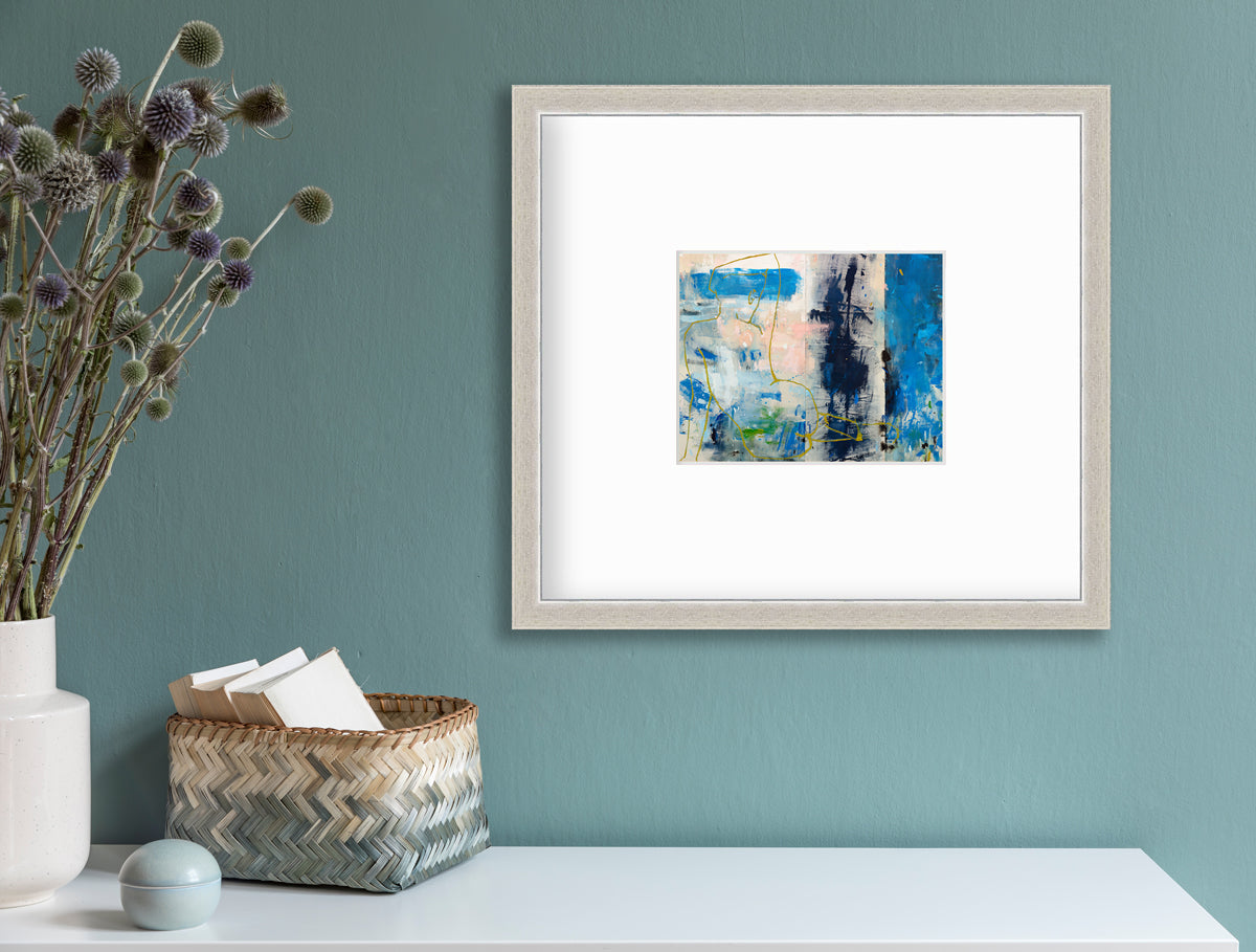 A blue, beige, gold and pink abstract figurative print in a silver frame with a mat hangs on a teal wall above a white surface with a beige and teal basket holding card stock, a round teal trinket and a white vase with green and purple pom pom stems.