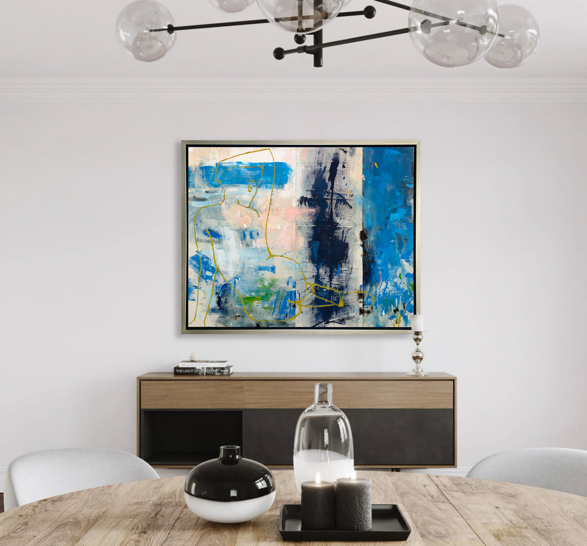 A blue, beige, gold and pink abstract figurative print in a silver floater frame hangs on a white wall above a wood and black sideboard with a stack of books, a candle and across from a round wood table with vases, candles and two white chairs.