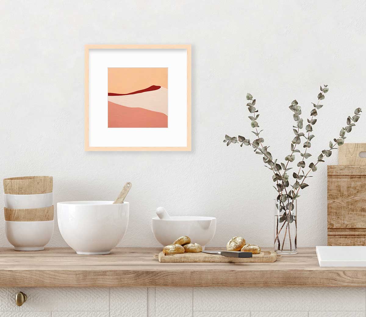 A red, peach and beige abstract print in a natural wood frame with a mat hangs on a white wall above a butcher block counter with bowls, a cutting board and a clear glass vase with eucalyptus stems.
