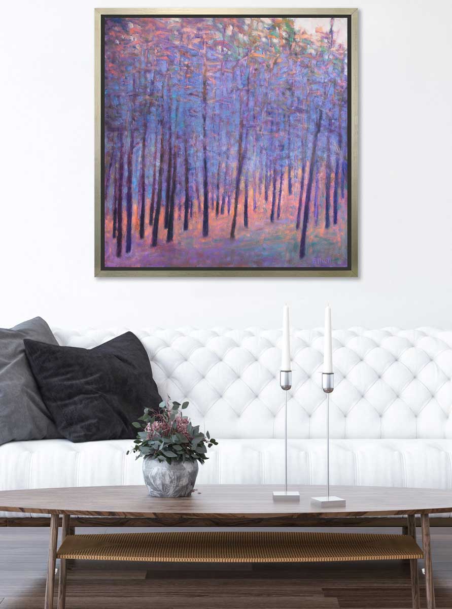 A blue and pink abstract tree landscape print in a silver floater frame hangs on a white wall above a white tufted sofa with grey and black pillows, a wooden oval coffee table, a small potted floral arrangement and two tall candlestick holders with white candles.