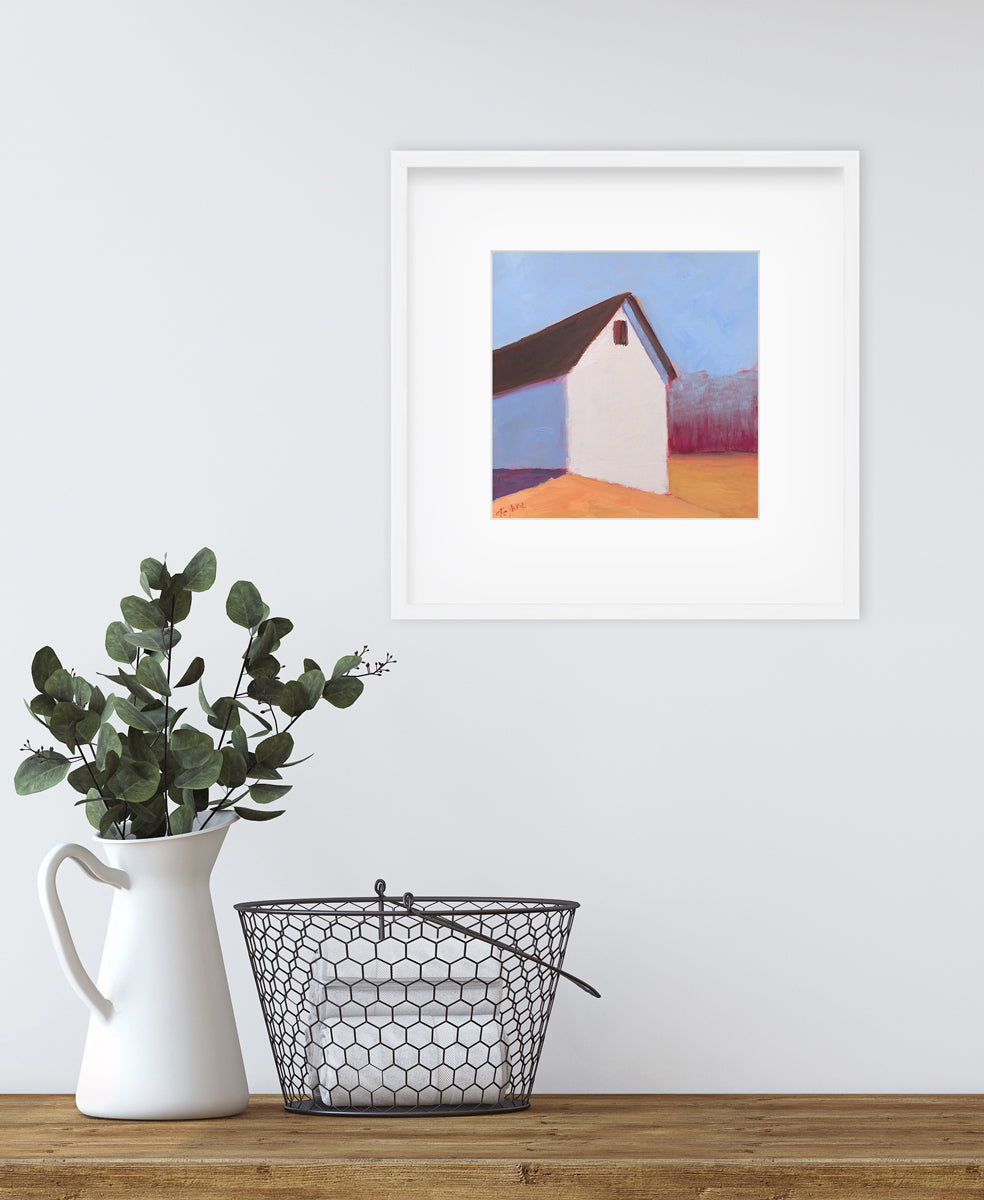 A blue, white, red and orange contemporary barn print in a white frame with a mat hangs on a white wall above a wood counter with a white vase holding eucalyptus and a metal basket sitting on top. 