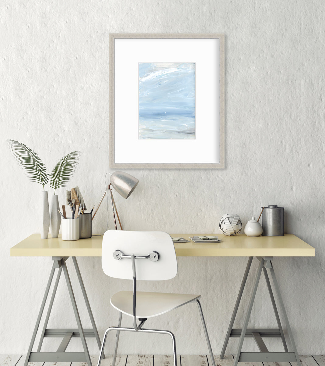 A white, blue and grey abstract seascape print in a silver frame with a mat hangs on a light grey wall above a natural wood and metal desk with decorative accents, a small chrome adjustable lamp and a white chair.