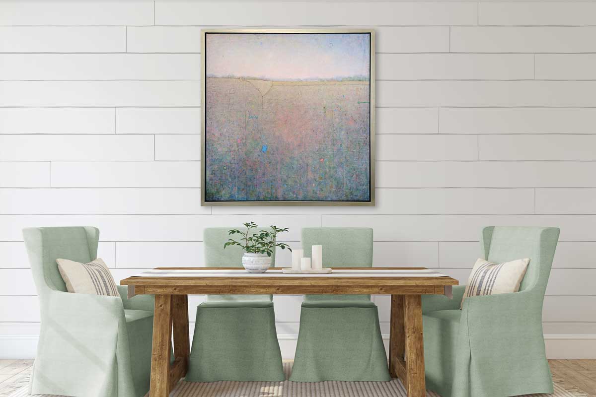 A green, pink and blue abstract landscape print in a silver floater frame hangs on a white shiplap wall above a wood dining table with four light green slip-covered chairs surrounding.