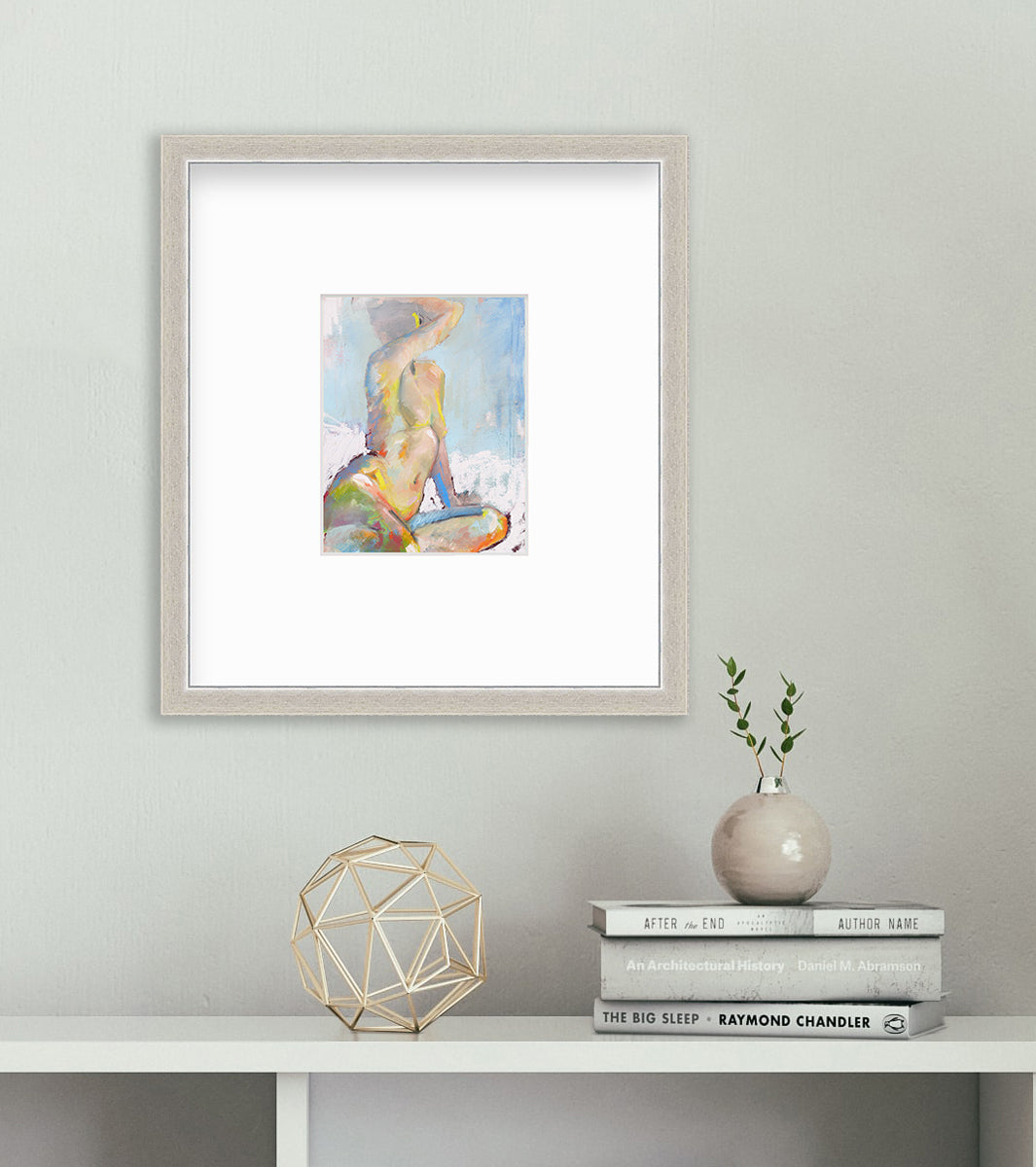 A multicolored abstract figural print in a silver frame with a mat hangs on a white wall above a white surface with a stack of books, a miniature vase with two small stems and a gold wire sphere decoration.