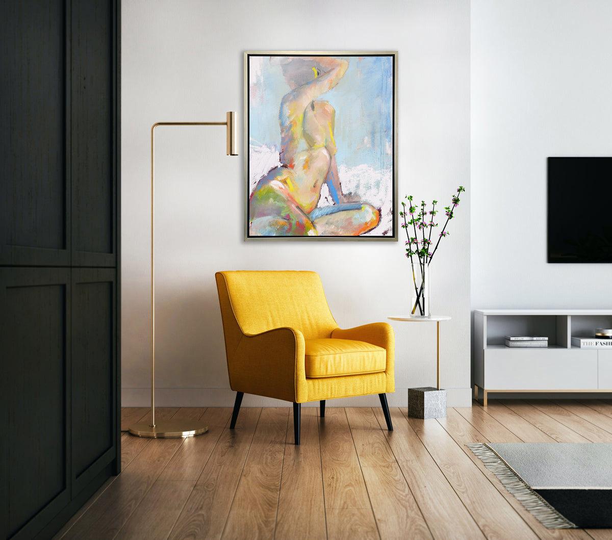 A multicolored abstract figural print in a silver floater frame hangs on a white wall above a dark yellow chair, a white marble side table with floral stems in a clear vase and a tall gold floor lamp.