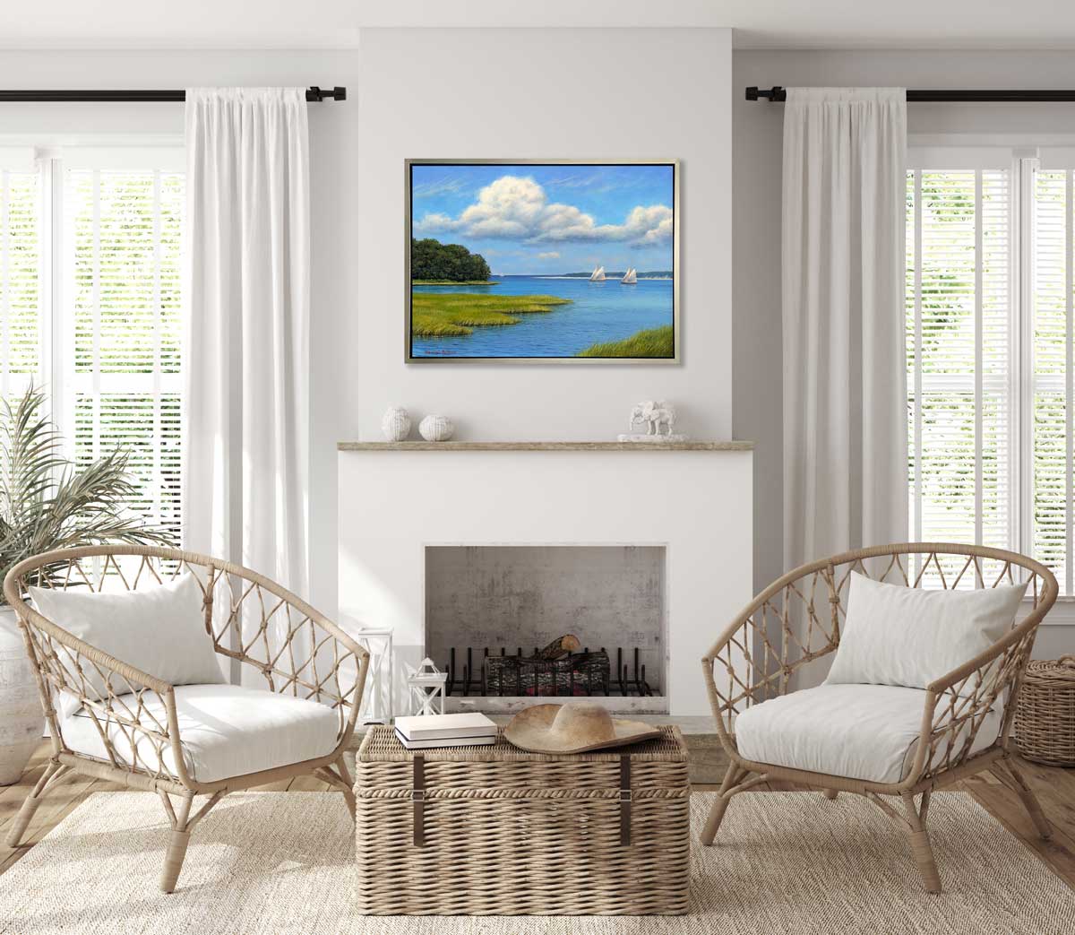 A realist contemporary seascape print with sailboats in a silver floater frame hangs on a white wall above a fireplace mantle, two woven chairs with white cushions and a wicker trunk.