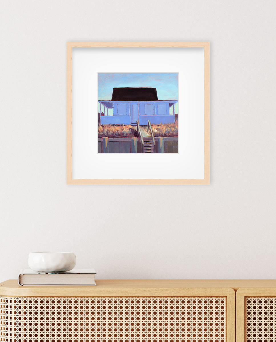 A light blue, lavender, burnt orange and brown contemporary cottage print in a natural wood frame with a frame hangs on a white wall above a beige wicker console with a book and white bowl sitting on top.  