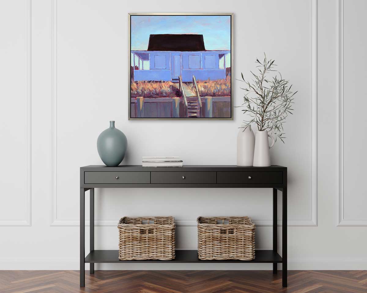 A light blue, lavender, burnt orange and brown contemporary cottage print in a silver floater frame hangs on a white wall above a black console table with a muted teal vase, two white vases with greenery and two wicker baskets.