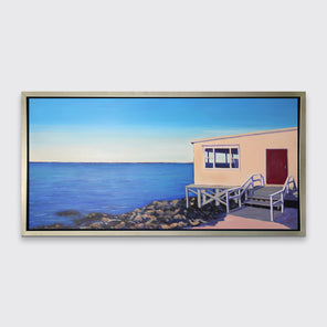A blue, beige and grey contemporary seaside print with a beach house on the rocks next to the ocean in a silver floater frame hangs on a white wall.