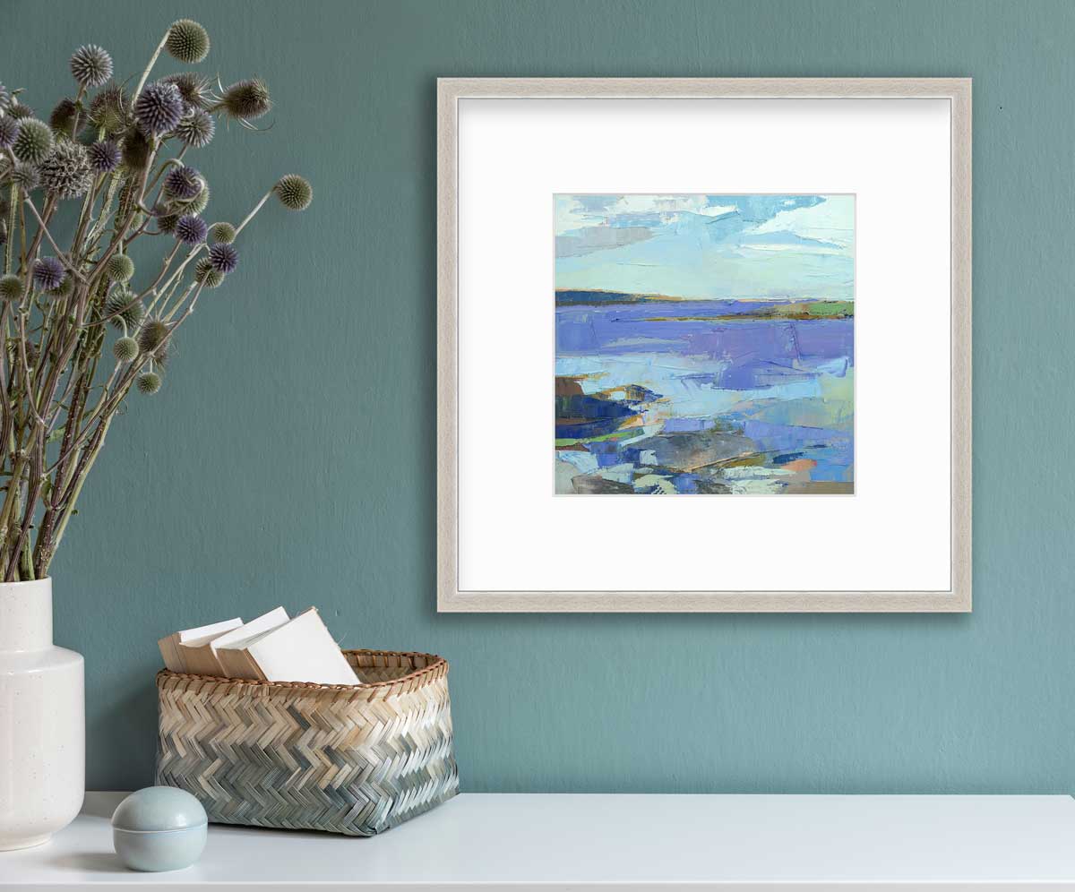 A blue, purple, white and green abstract landscape print in a silver frame with a mat hangs on a teal wall above a white surface with a beige and teal woven basket full of card stock, a light teal round trinket and a white vase with green and purple pom pom stems.