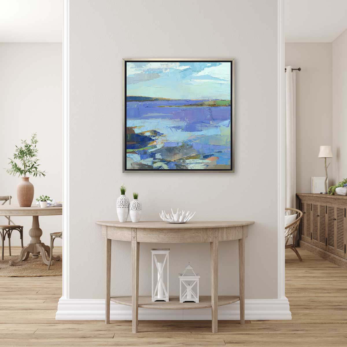 A blue, purple, white and green abstract landscape print in a silver floater frame hangs on a white wall above a wood half-moon console table with two white vases, a white decorative bowl and two white lanterns.