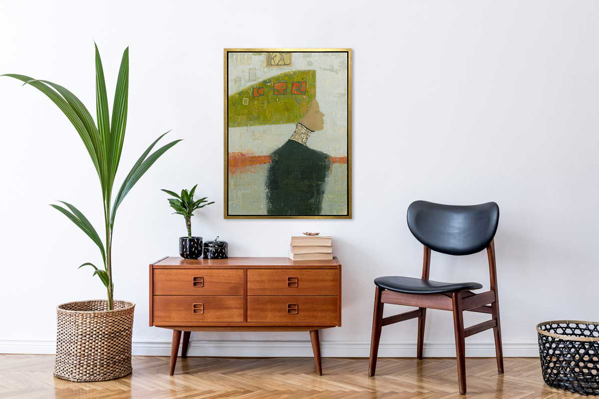A grey, green and orange abstract print of a woman with a headpiece in a gold floater frame hangs on a white wall above a wood cabinet with a stack of books and a small potted plant, a black chair and a potted floor plant.
